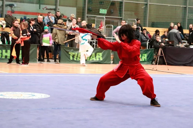 Kobieta w czerwonym kimono ustawia się w pozycji bojowej. Jest skupiona. W prawej ręce trzyma miecz, który jest skierowany w stronę potencjalnego przeciwnika