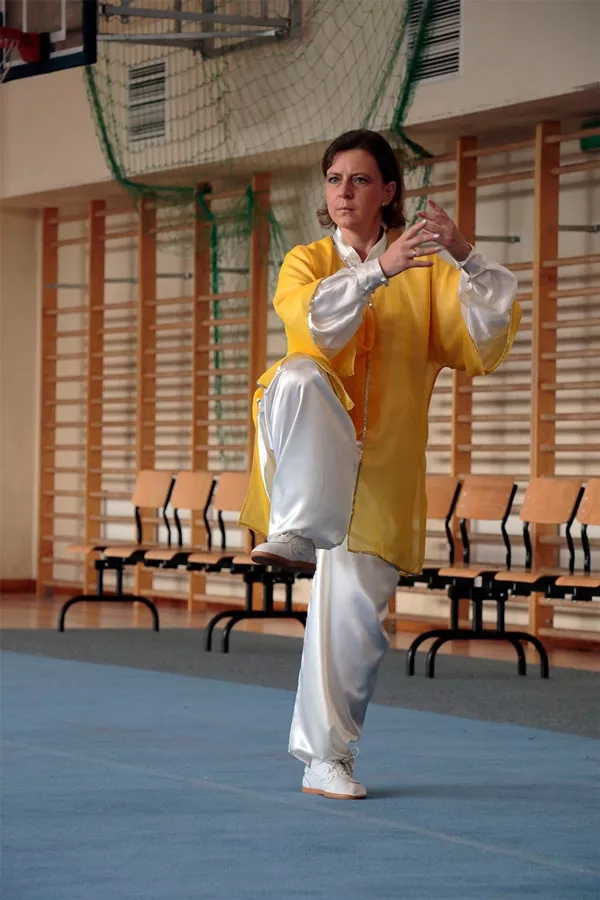 Kobieta w biało-żółtym stroju utrzymuje równowagę stojąc na lewej nodze, a prawą trzymając w górze. Jej ręcę są ułożone w koszyczek, jakby miała w nim się mieścić energia