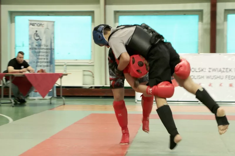 Zawodnik w czerwonym kasku chwyta w kolanach zawodnika w czarnym kasku i próbuje go przewalić za siebie.